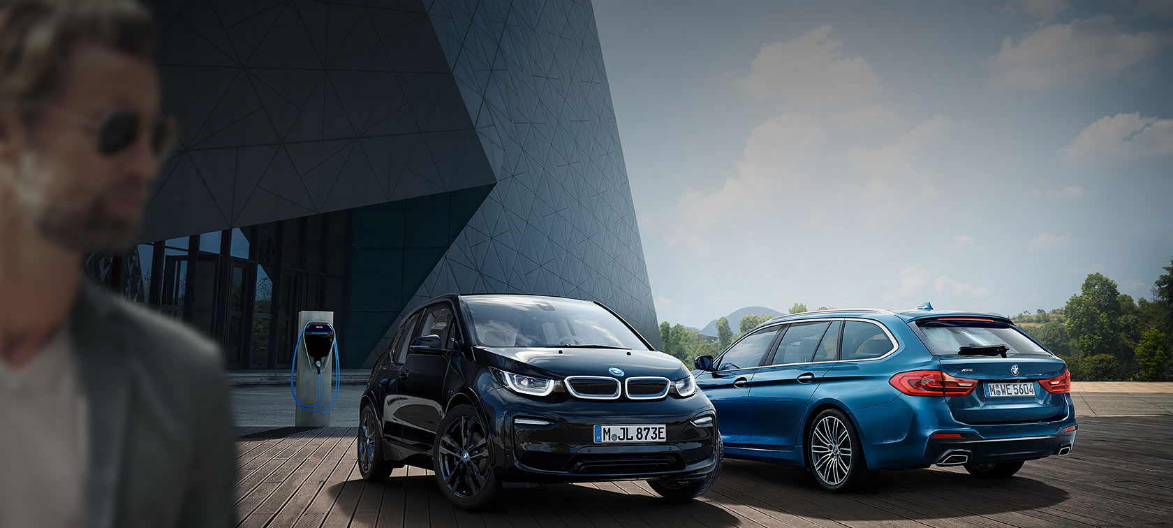 Zwei BMW-Autos, ein schwarzer i3 und ein blauer Kombi, parken vor einem modernen Gebäude.