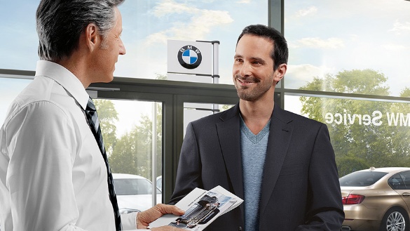 Zwei Männer in einem BMW Autohaus, einer hält Prospekte, im Hintergrund Autos und BMW-Logo.