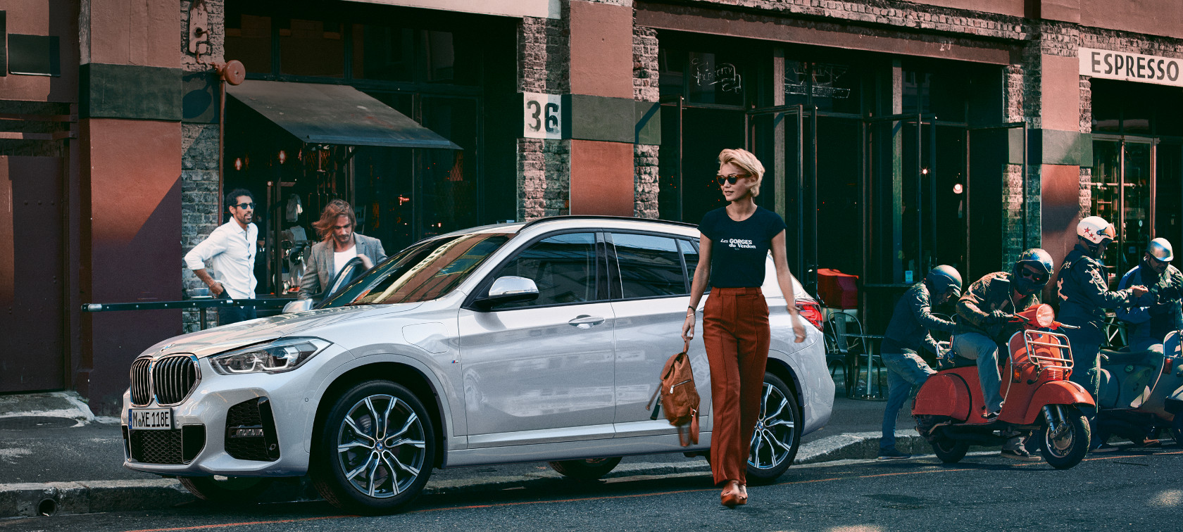 Silberner BMW X1 steht am Straßenrand vor einem Café, mehrere Personen und rote Motorroller.