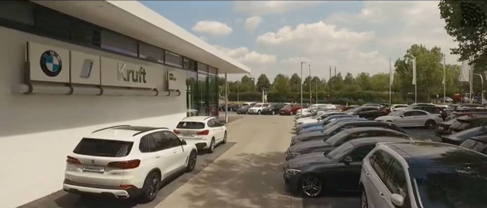 BMW Autohaus mit geparkten Autos, Gebäude mit Kruft-Logo, blauer Himmel, Bäume im Hintergrund.