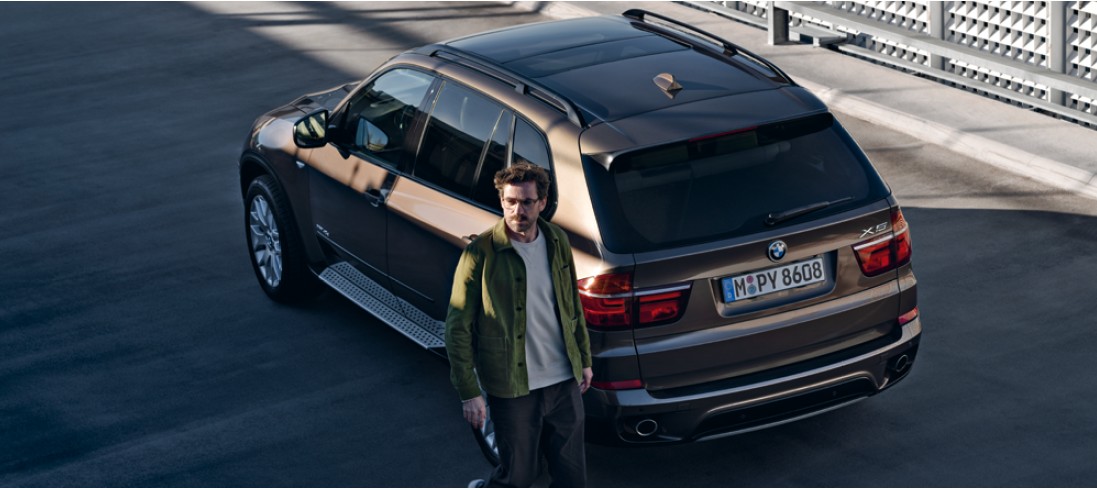 Mann in grüner Jacke steht neben einem braunen BMW X5 auf einem Parkplatz.