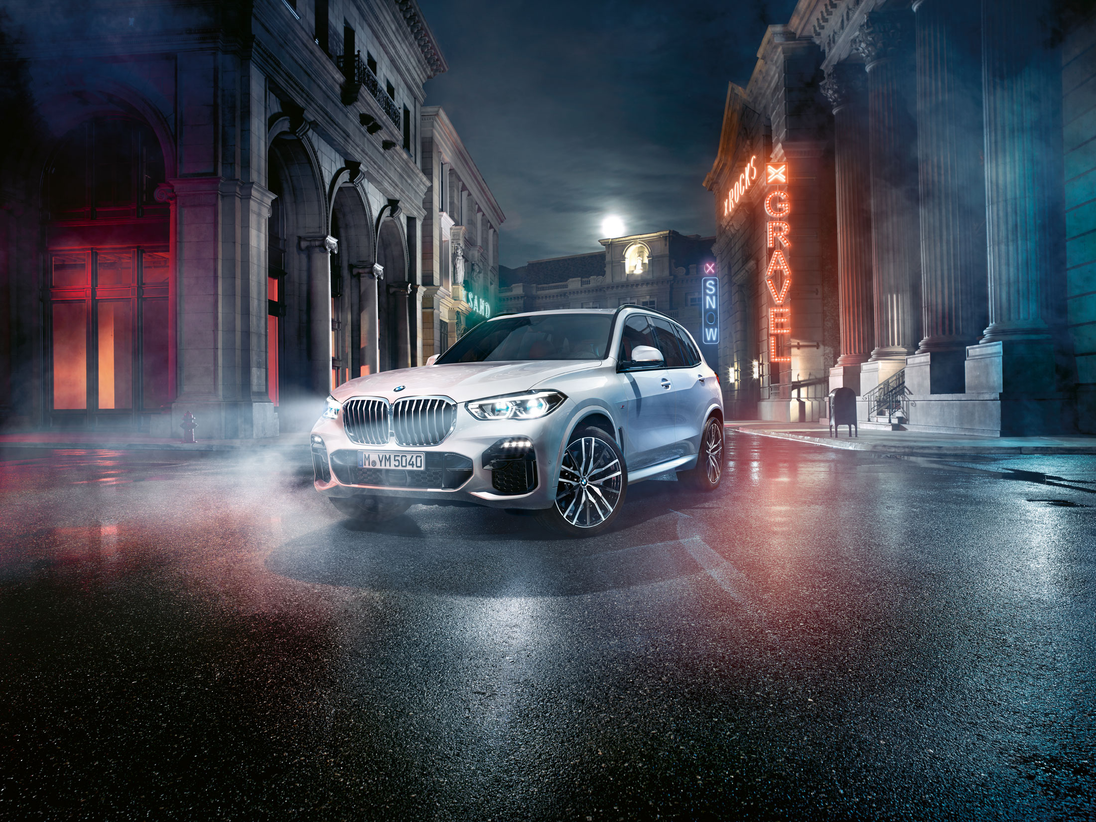 Silberner BMW X5 auf nasser Straße bei Nacht, umgeben von neoklassischen Gebäuden und Neonlichtern.