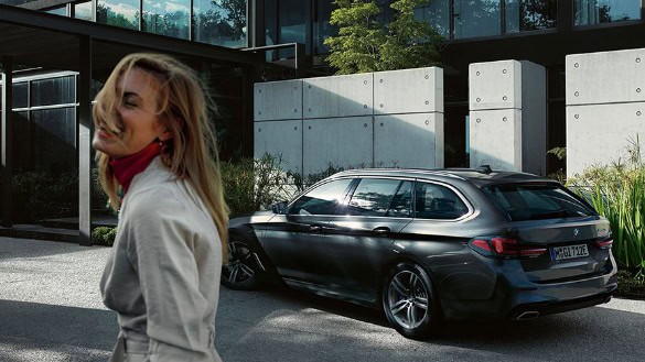 Frau lächelt vor grauem BMW Kombi auf Parkplatz, moderne Architektur im Hintergrund.