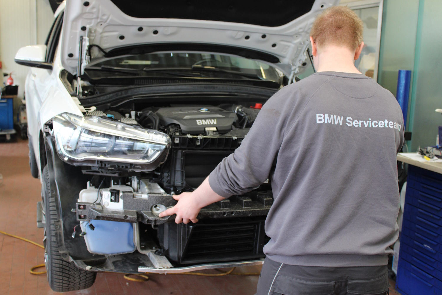 Eine Person im BMW Serviceteam-Pullover arbeitet an einem zerlegten weißen BMW mit geöffneter Motorhaube.