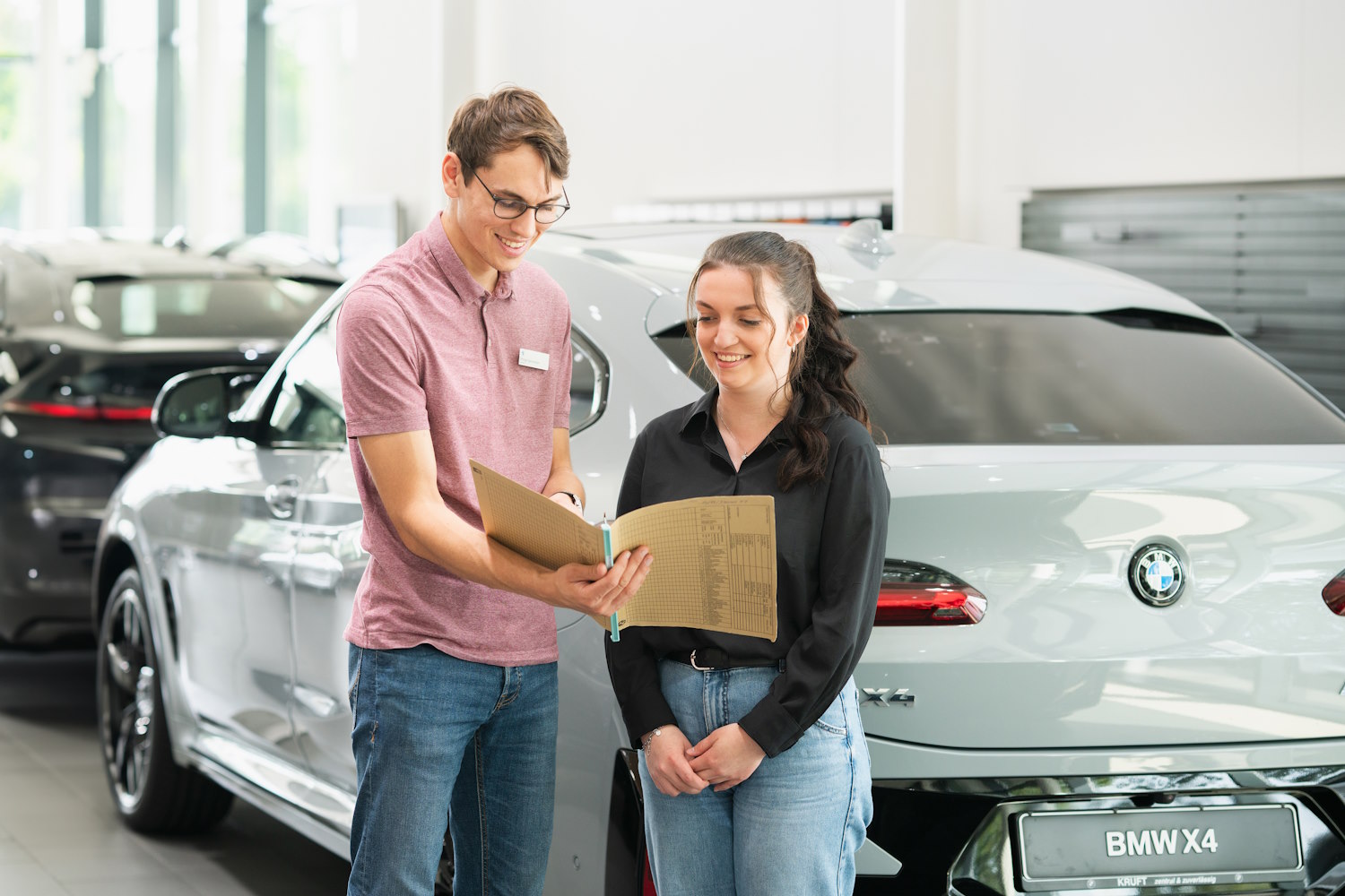 Zwei Personen vor einem silbernen BMW X4 im Autohaus, eine Person hält eine Mappe.