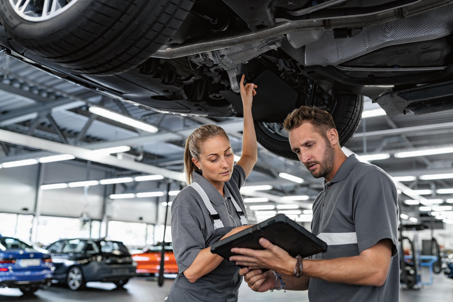 Zwei Techniker in grauer Arbeitskleidung inspizieren ein Auto in einer Werkstatt.