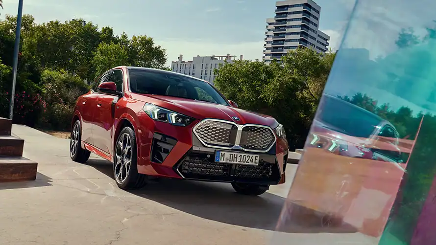Roter BMW iX2 steht auf einer Straße, im Hintergrund sind Bäume und moderne Gebäude zu sehen.