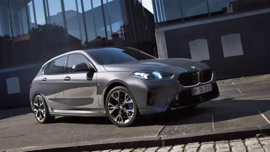 Grauer BMW 1er parkt vor einem modernen Gebäude mit Glasfassade auf einer gepflasterten Straße.