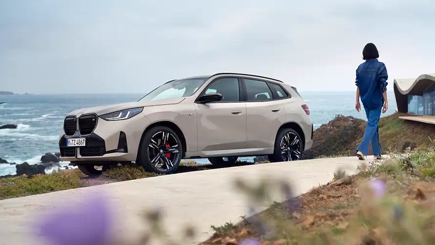 BMW X3 M50 in Beige steht an einer Küstenstraße, eine Person geht zu einem Gebäude.
