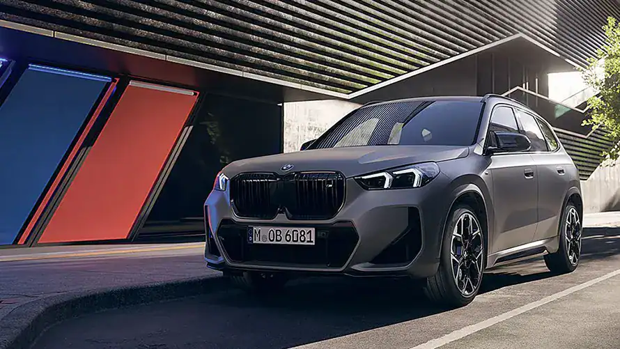 Grauer BMW-SUV vor moderner Architektur mit roten und blauen Akzenten, seitliche Perspektive.