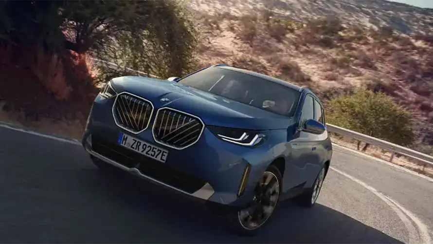 Blauer BMW X3 fährt auf kurviger Straße, umgeben von Bäumen und Hügeln.