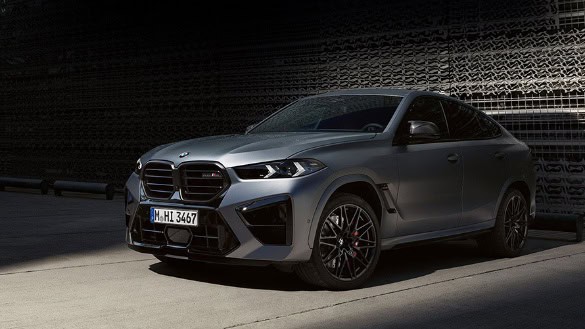 Silberner BMW X6 M auf dunkler Straße, seitliche Frontansicht, Schatten im Hintergrund.